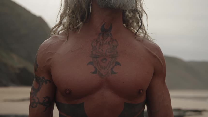 The video is a closeup shot of a shirtless man with long wavy blondegray hair and a beard standing outdoorshe is heavily tattooed on his chest and arms with dark tribalstyle designs and is looking up and away from the camera in a dramatic
