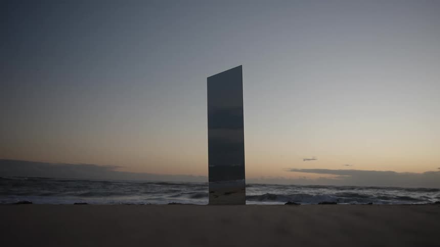 Tall rectangular mirror standing on the sand reflects the sea and the sky at dusk