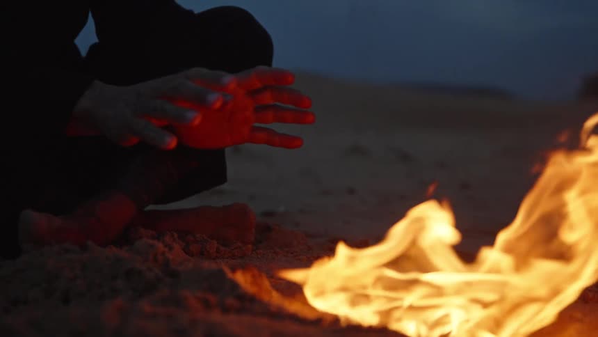 A person sits on the ground near a patch of bright fire extends hands toward the heat rubs hands together and then raises the head to show a face with a beard and hair pulled back against a dark sky
