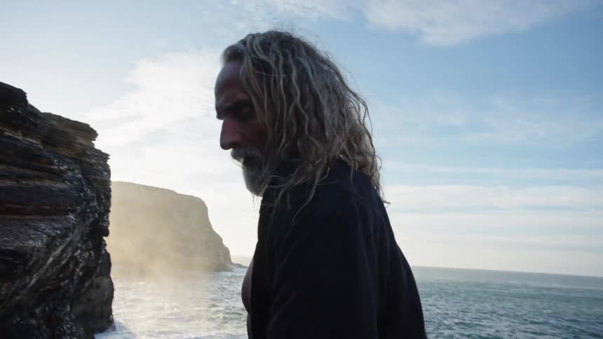 An old man with long hair and a beard dancing peculiarly on rocks near the sea