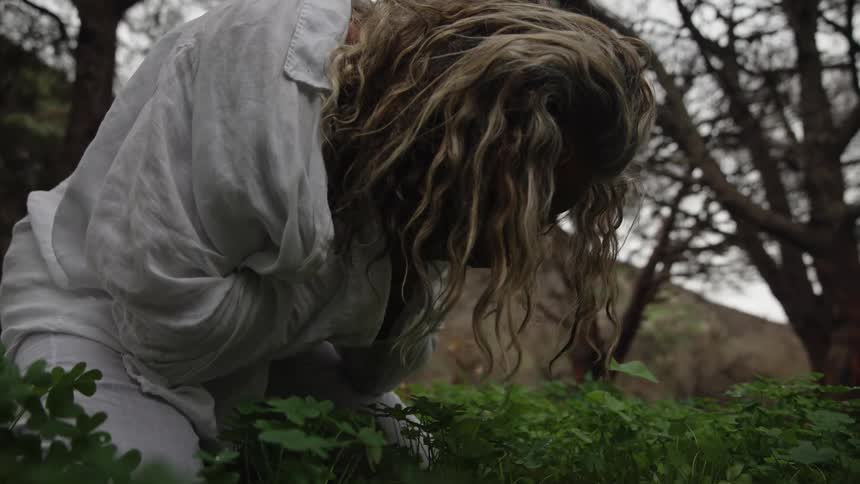 A person with long wavy hair in a loose white garment bends over green groundcover outdoors appearing somber
