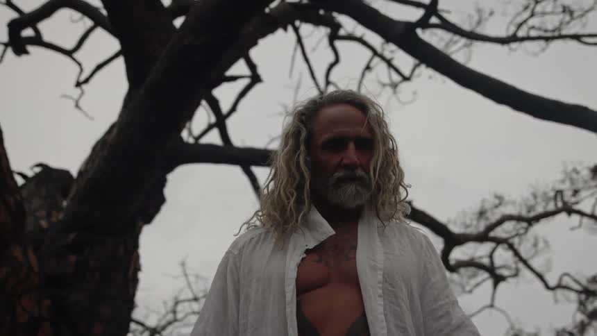 A muscular man with long blonde hair and a beard wearing an open white shirt stands looking forward with an intense expression beneath the dark silhouetted branches of a tree against an overcast sky