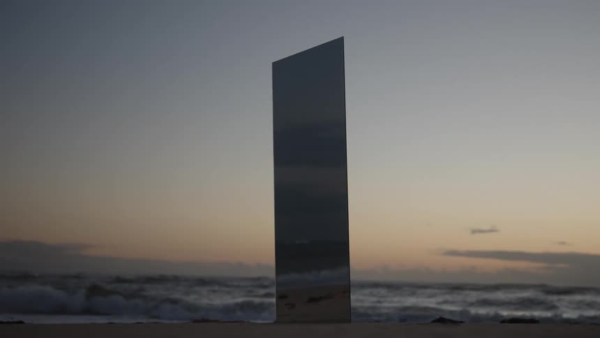 A sleek reflective mirror stands vertically on a dark beach with the ocean waves breaking behind it under a soft twilight sky