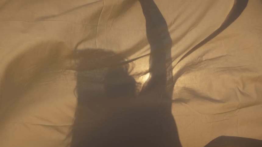 Shadow of person with long hair and raised arm is cast on fabric lit by warm light