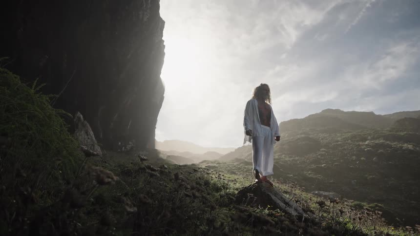 The video shows a person in a flowing white outfit standing on a dark rocky outcrop looking out over a misty mountainous landscape the scene is brightly backlit by a strong sun or spotlight creating a dramatic ethereal silhouette against the l