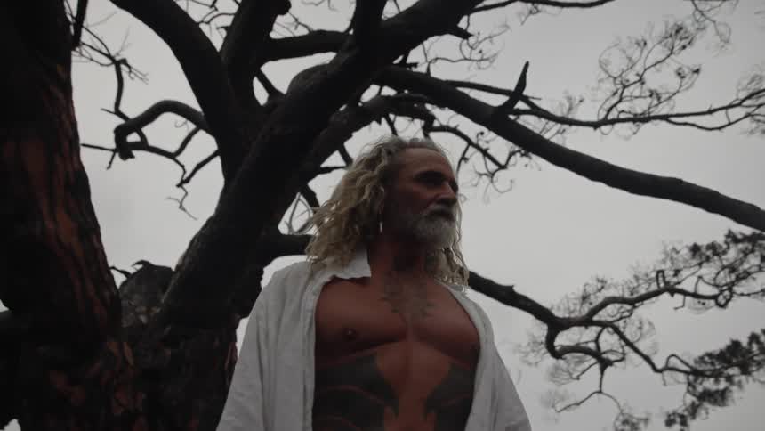 Tattooed man with light hair standing in front of leafless tree under cloudy sky