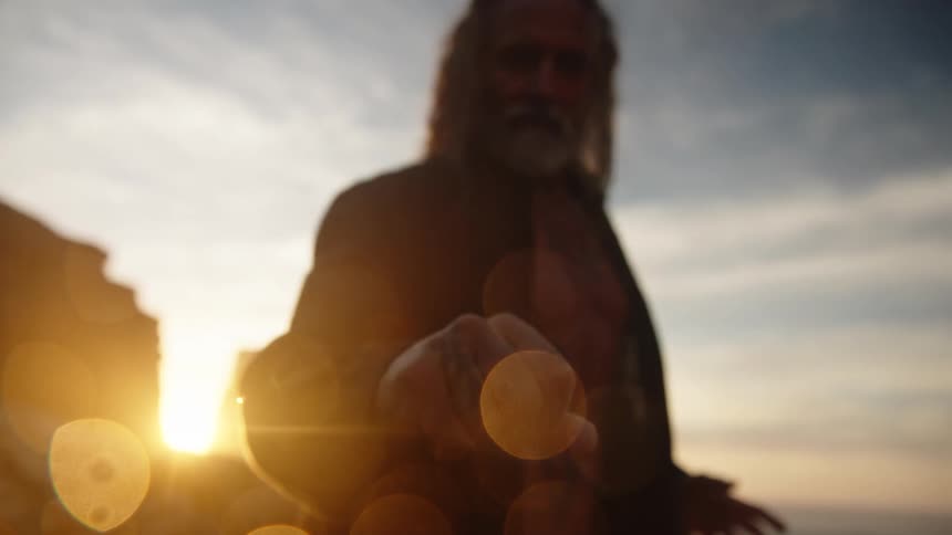 An elderly man extends one hand toward the camera lens the bright sun sits low in the distance behind the person light creates flares and large round bokeh shapes all in a coastal setting