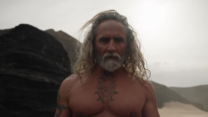 A close view shows a shirtless older man with long blonde hair a gray beard and a chest tattoo looking directly at the camera on a dark rocky beach under an overcast sky