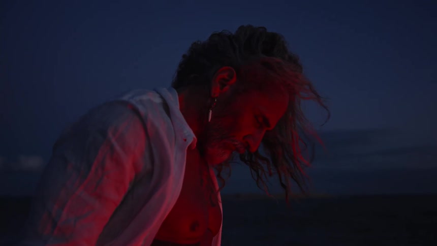 A side close view of a shirtless older man with long hair a gray beard and silver earrings wearing an open white shirt illuminated by a red light against a dark blue background