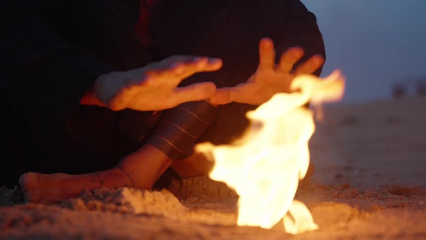 The man warms his hands on the campfire on the beach
