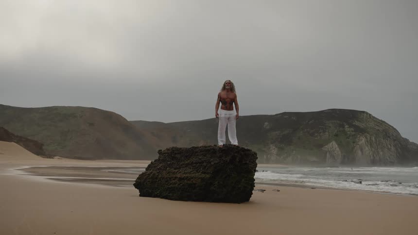 A shirtless middleaged man stands on a rock at the beach on a cloudy day