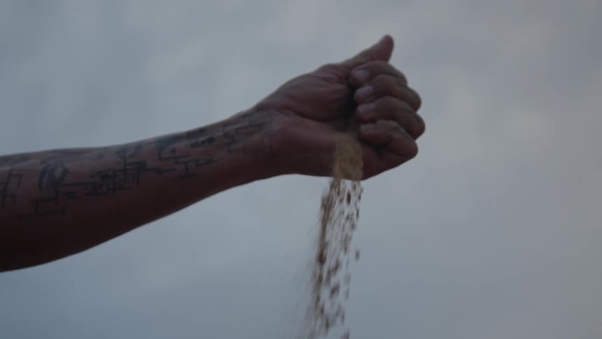 A persons tattooed forearm and closed hand extend into the air a stream of granular material falls from the fingers and the background is a overcast sky