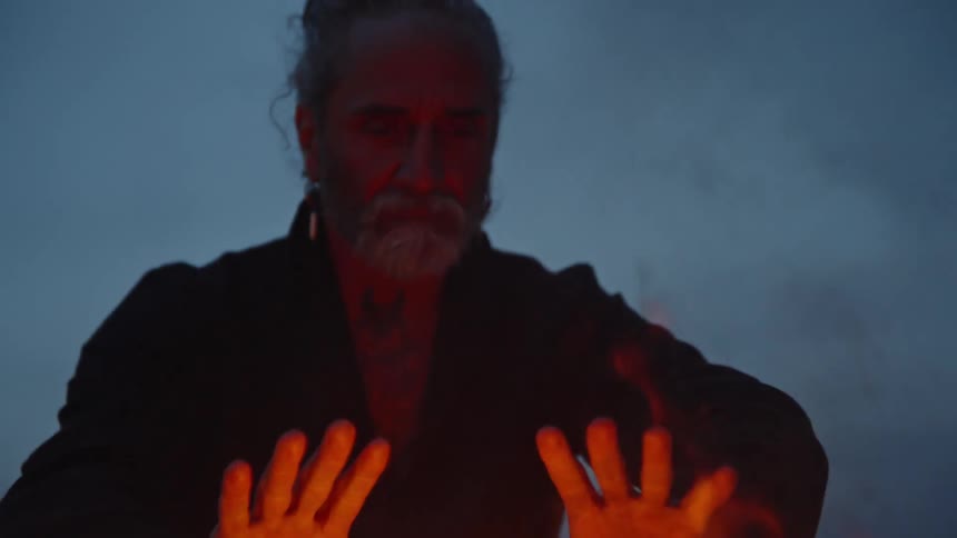 Older man holds his hands close to the campfire while sitting outdoors under an overcast sky