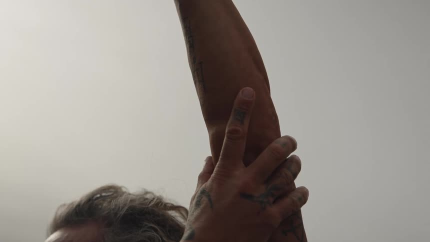 Man with visible tattoos on raised arm and overcast sky in the background