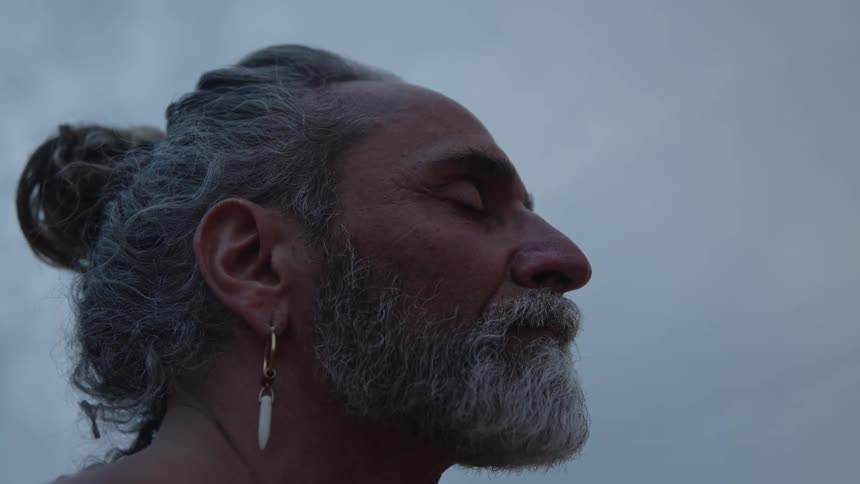 A close side view shows an older man with gray hair in a bun a white beard and a tattooed upper body wearing a hoop earring against a dim overcast sky