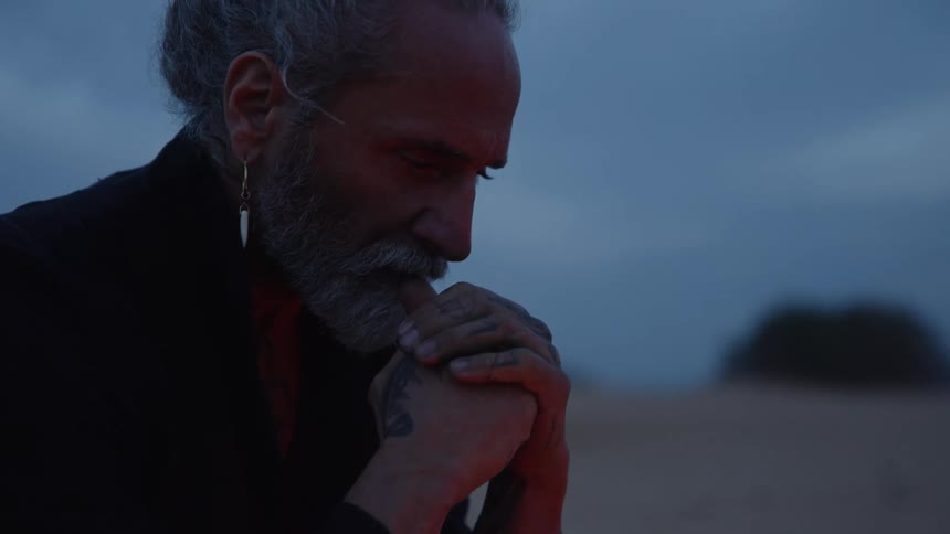 The video features an extreme closeup of a man with a white beard and a top knot looking down with the scene lit by a dramatic red and blue light he appears to be seated or kneeling outdoors at twilight or dawn possibly in a wide desolate are