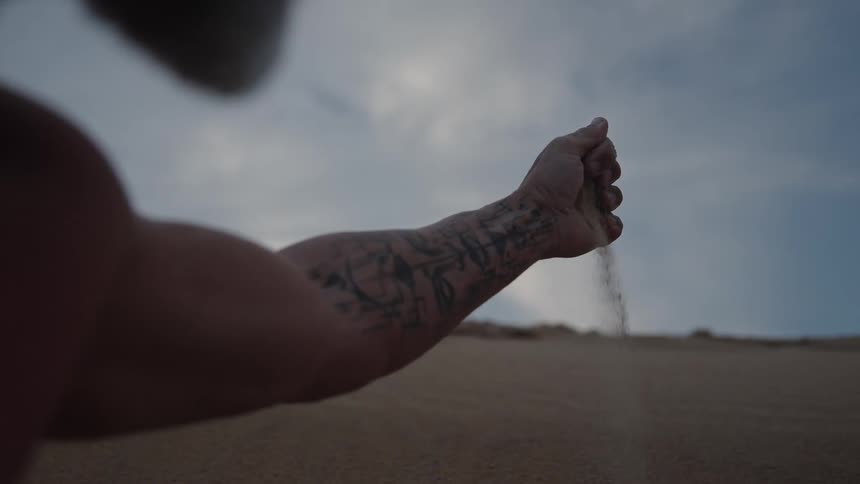 A persons tattooed arm and hand the person holds a handful of sand and allows it to trickle downward and the dark figure is silhouetted against a cloudy sky over a vast sandy landscape