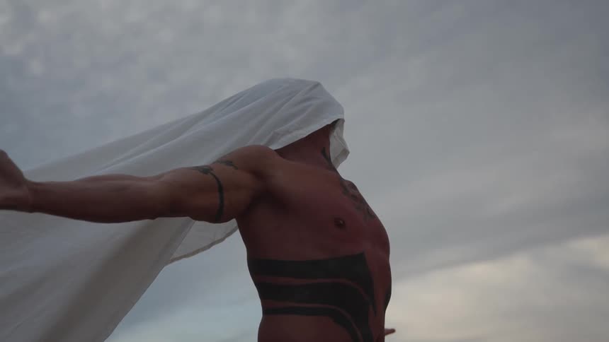 A shirtless man stands outdoors at midday beneath a cloudy sky his face covered with a white cloth and his body marked with multiple tattoos