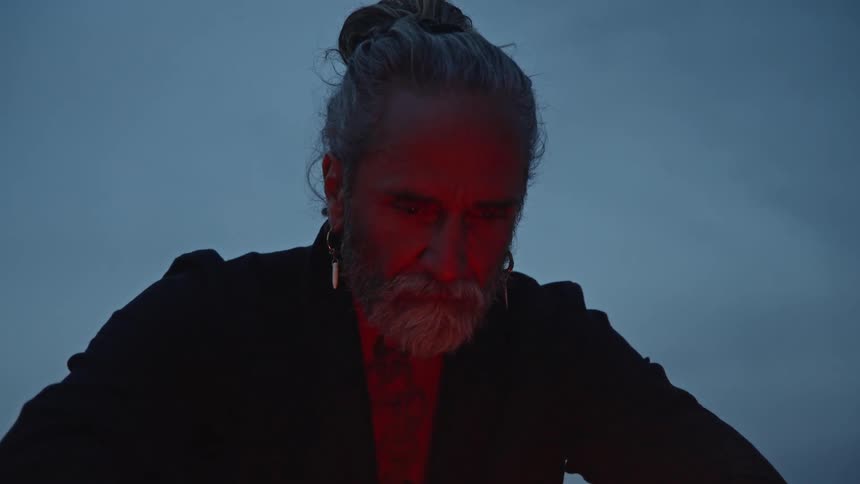 A medium closeup shows a man with a long gray beard and hair tied in a top knot looking down his face partially illuminated by the orange glow of a small fire in the foreground he is dressed in dark clothing and the background is a dim twi