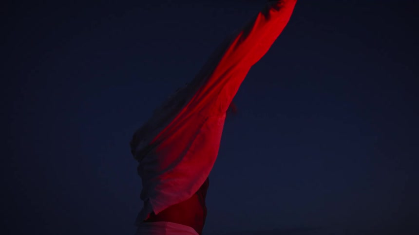A medium view shows a shirtless bearded man with long hair covered in red light standing arms wide open against a dark blue sky while wrapping himself in a flowing white shirt
