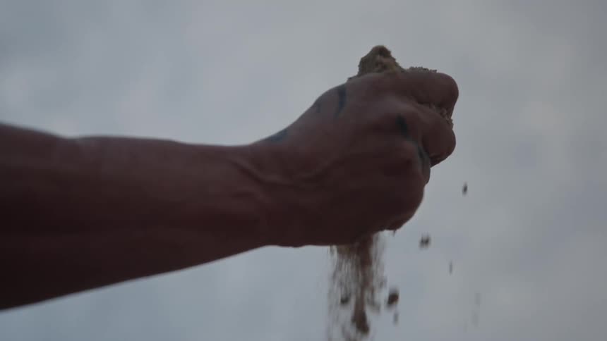 Tattooed hand holding sand with particles falling against cloudy sky