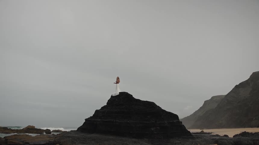 Person in white dress stands on dark rock at beach with overcast sky and ocean in the background