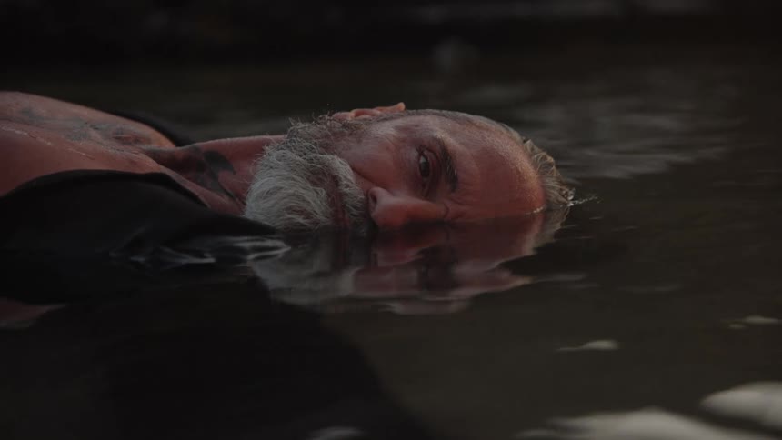 A middleaged man with a gray beard lies motionless in shallow water at midday