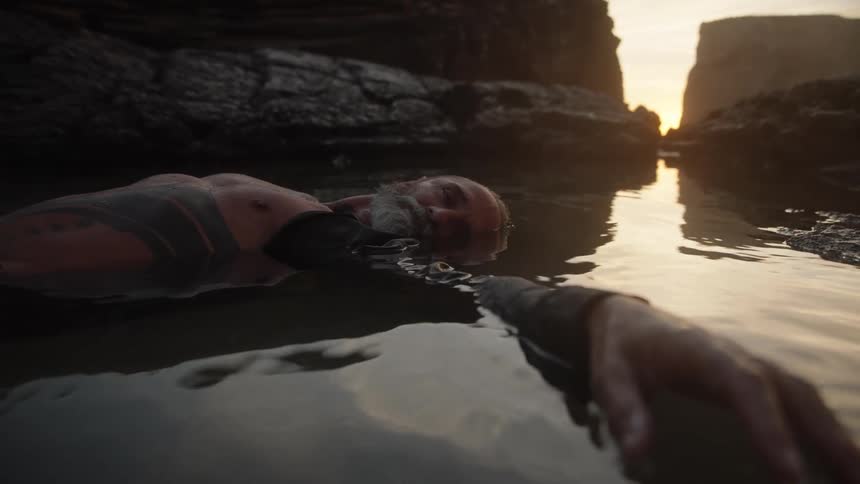 Older man lies in water reflecting the red sky and seaside cliffs