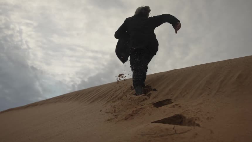 Person dressed in black runs uphill on a steep sand dune under a cloudy sky