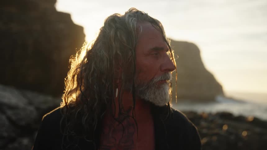 A closeup shows an older man with long backlit hair and a gray beard standing on a rocky coastline at sunset looking downward before lifting his head and turning his face toward the light