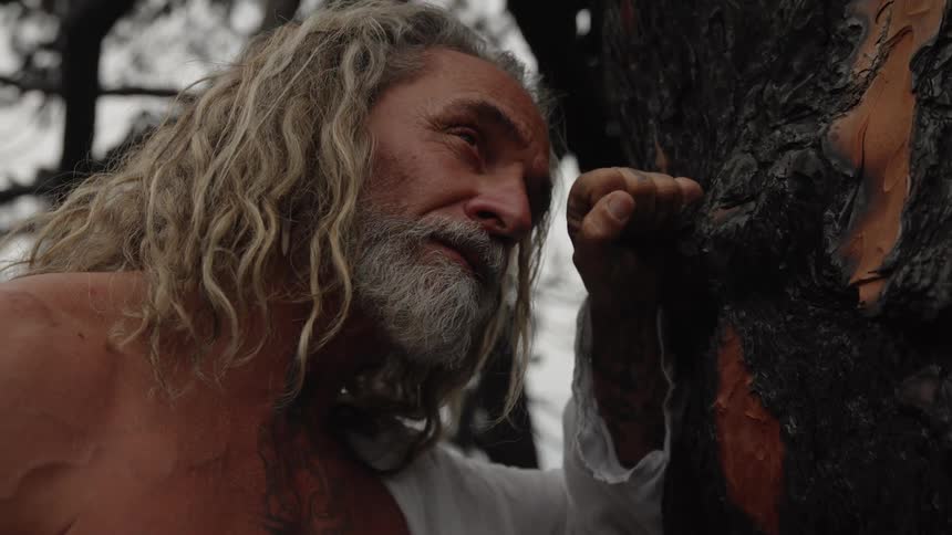 A close view shows a shirtless tattooed older man with long blonde hair and a gray beard wearing a white shirt resting his forehead and fist against the dark cracked bark of a burned tree trunk