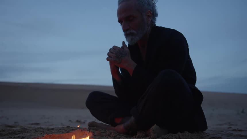 A middleaged man sits crosslegged on the beach at sunset warming himself by a campfire