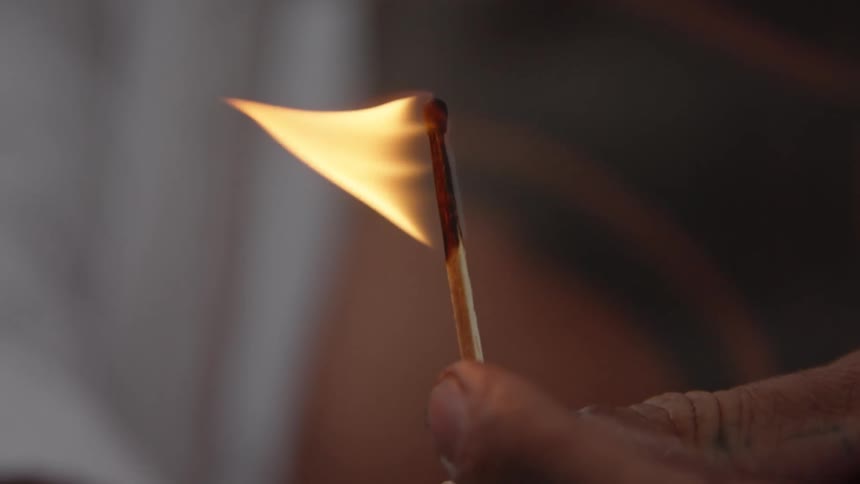 A close view shows a persons hand holding a wooden matchstick that is flaming brightly with a warm orange fire against an out of focus background