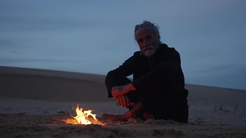 An old man sitting on the sand near a small campfire