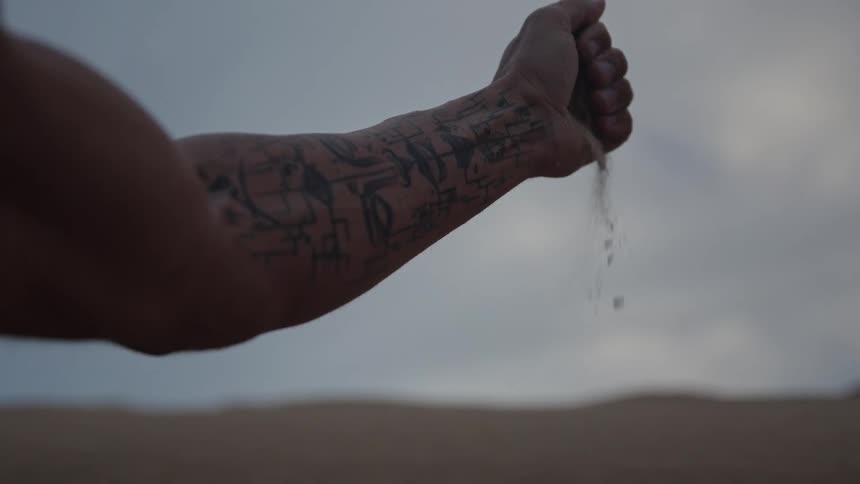 A tattooed arm and clenched hand are held horizontally against a bright overcast sky allowing sand to trickle slowly from the fist down to the dark ground below