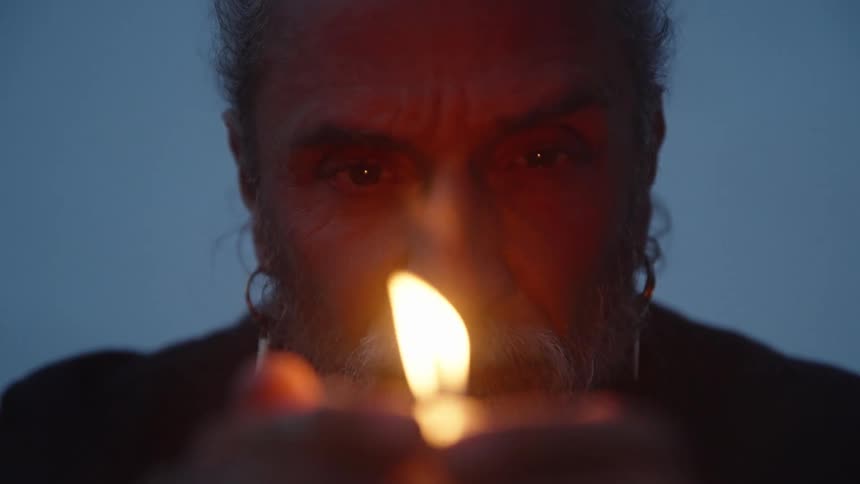 Older person with serious expression holding lit flame against dark background