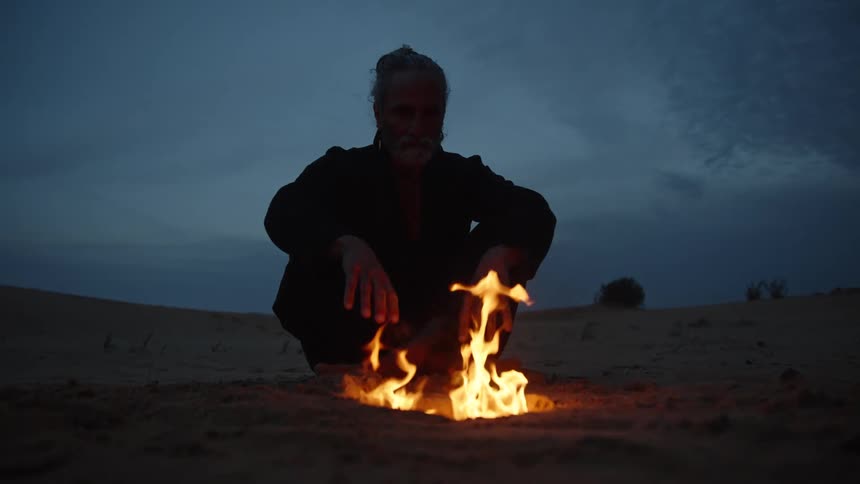 The man sits by the campfire in the sand as the sun sets