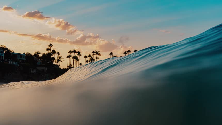 A large ocean wave begins to crest captured from a low water level with a silhouetted coastline of palm trees and houses visible under a colorful sunset sky