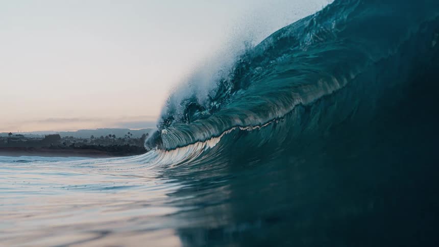 A large ocean wave curls and breaks forming a hollow tube of dark water with the shore and background hills visible under a fading sky