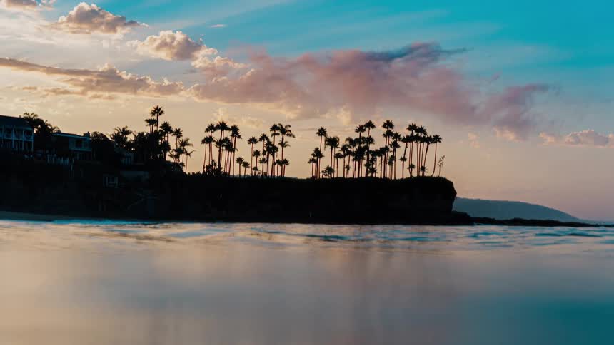 A silhouetted line of palm trees atop a seaside cliff with houses and hills in the background set against a colorful sky with the waters surface reflecting the scene in the foreground