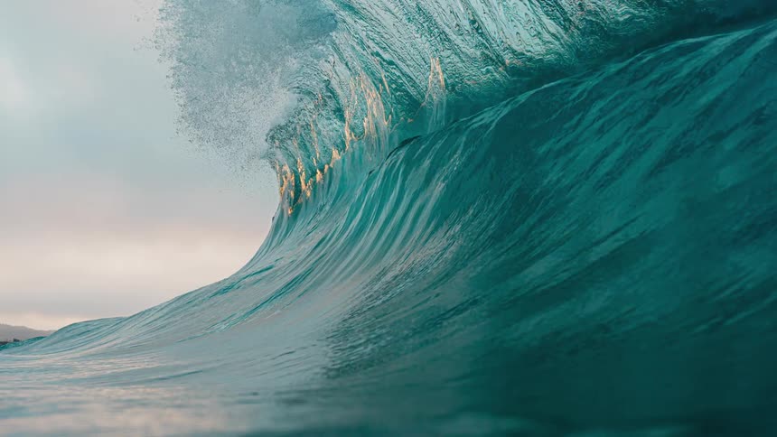 A low angle close view shows a massive powerful dark blue ocean wave cresting and beginning to break with white spray against a pale sunset sky