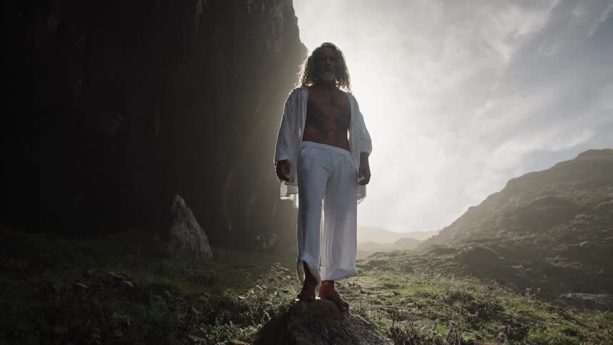 A middleaged man stands barefoot on a hill under the bright sun dressed in white clothing