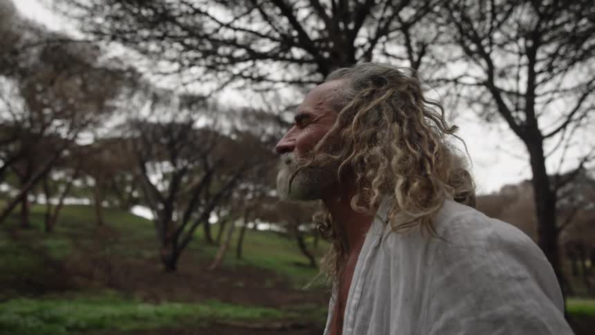 An old man with long hair kneeling on the ground among trees lamenting