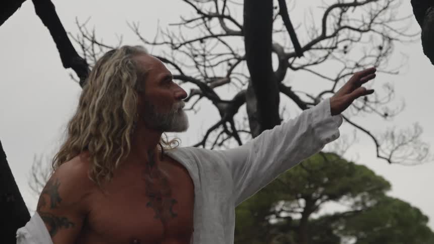 A man with long hair and tattoos stands among dark tree trunks touches the wood surface a white garment hangs around the mans shoulders and a field of grass and more trees fills the background