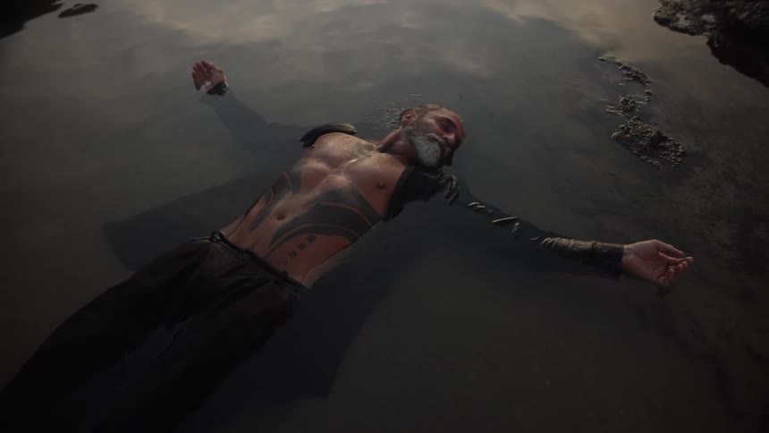 A middleaged man with a gray beard lies in shallow water at sunset his shirt unbuttoned to reveal tattoos on his body