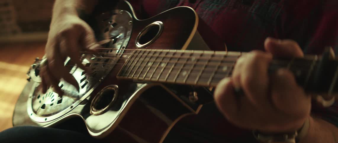A close view shows a person with rings on their fingers strumming a resonator guitar with a silver cover plate while wearing a red and black plaid shirt