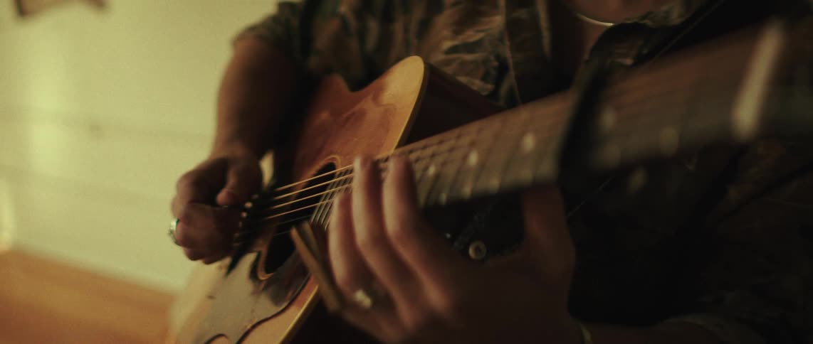 A close view of a person wearing a patterned shirt and rings on their fingers as they strum and finger chords on an acoustic wooden guitar in soft warm indoor lighting