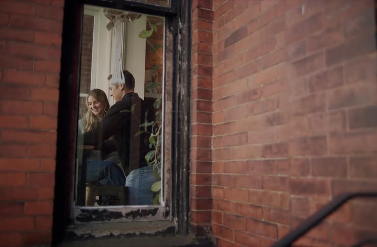 The video shows a smiling blonde woman sitting inside a room with her face visible through an open windowa man approaches the window and shakes the womans hand through the opening framed by the dark brick exterior of the building