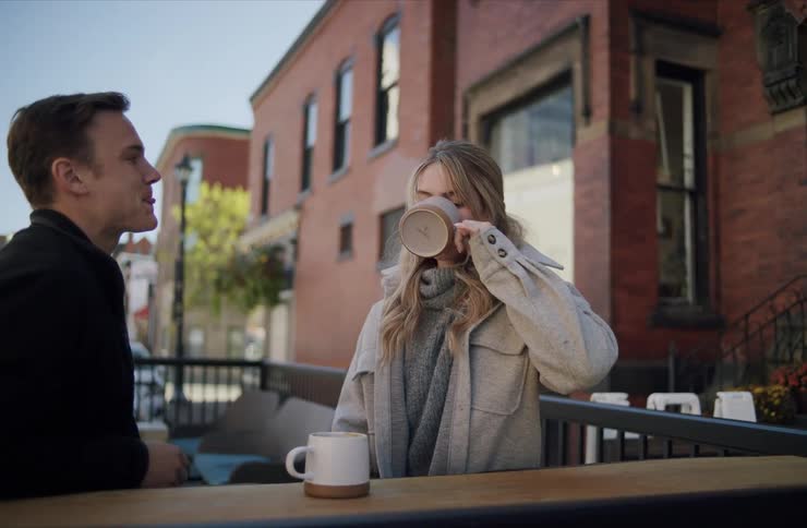 A man and a woman sitting across a wooden table rail outdoors each holding a beverage mug while the woman raises her mug to drink and the man looks at her in an urban setting with brick buildings in the background