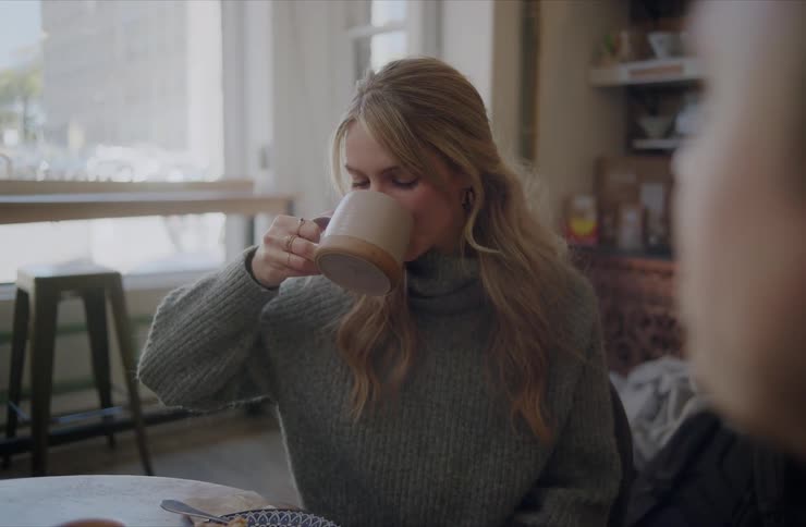A close view of a young woman with long blonde hair wearing a gray sweater as she sips from a coffee mug while sitting across from a person at a wooden table in a brightly lit cafe